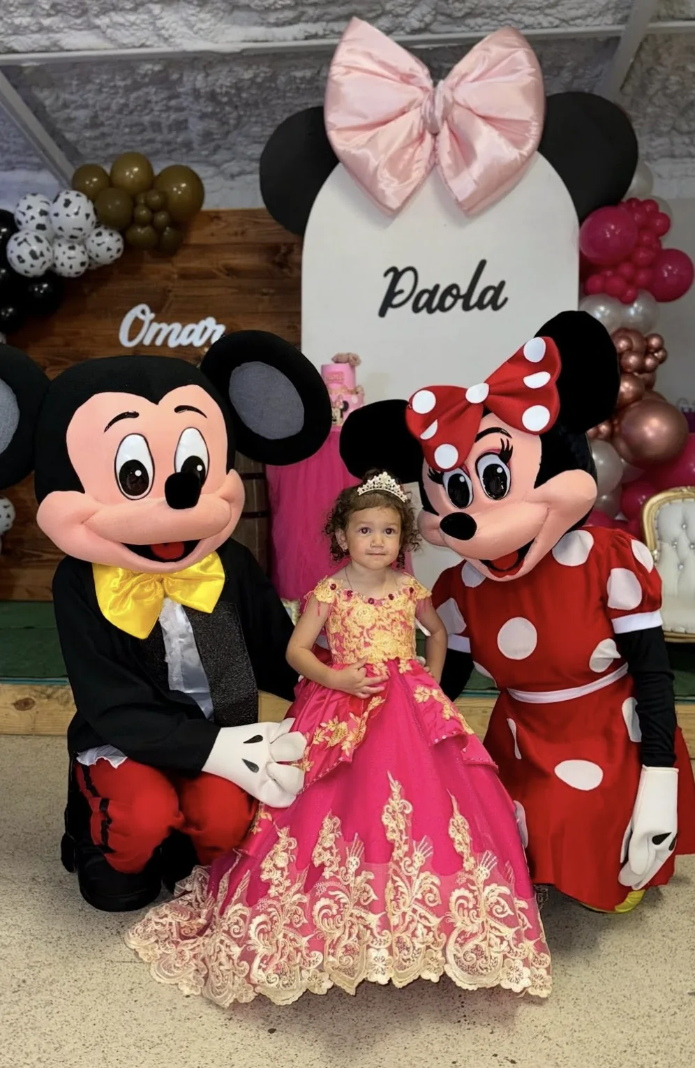 Child in a pink dress between Mickey and Minnie characters at a birthday celebration